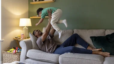 Man laying on a couch tossing his toddler son in the air and catching him.
